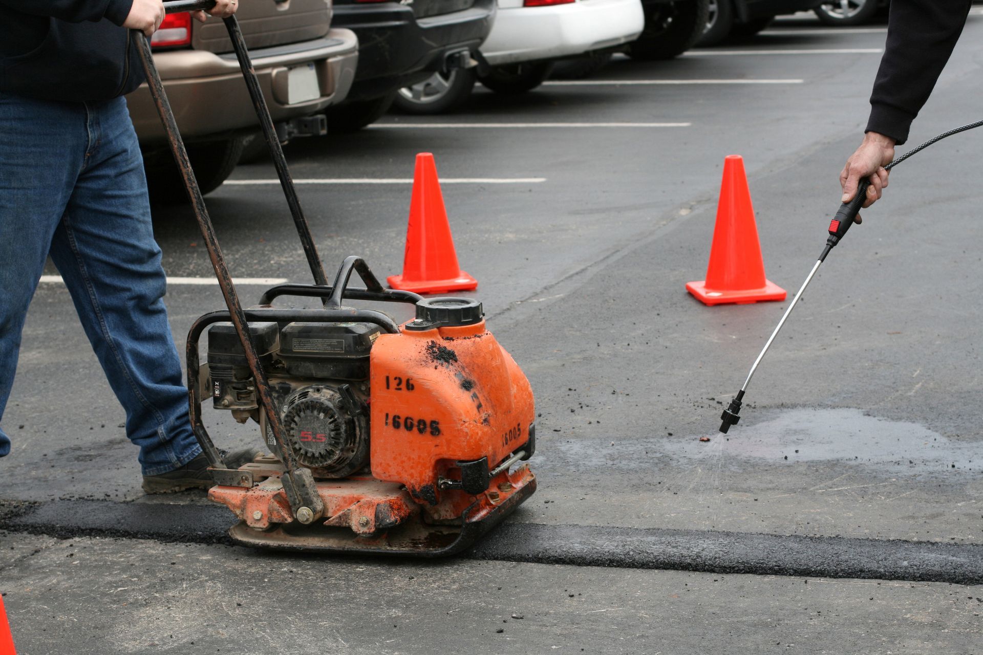 A construction worker shovels asphalt, wearing orange and blue reflective gear, by a paving machine.