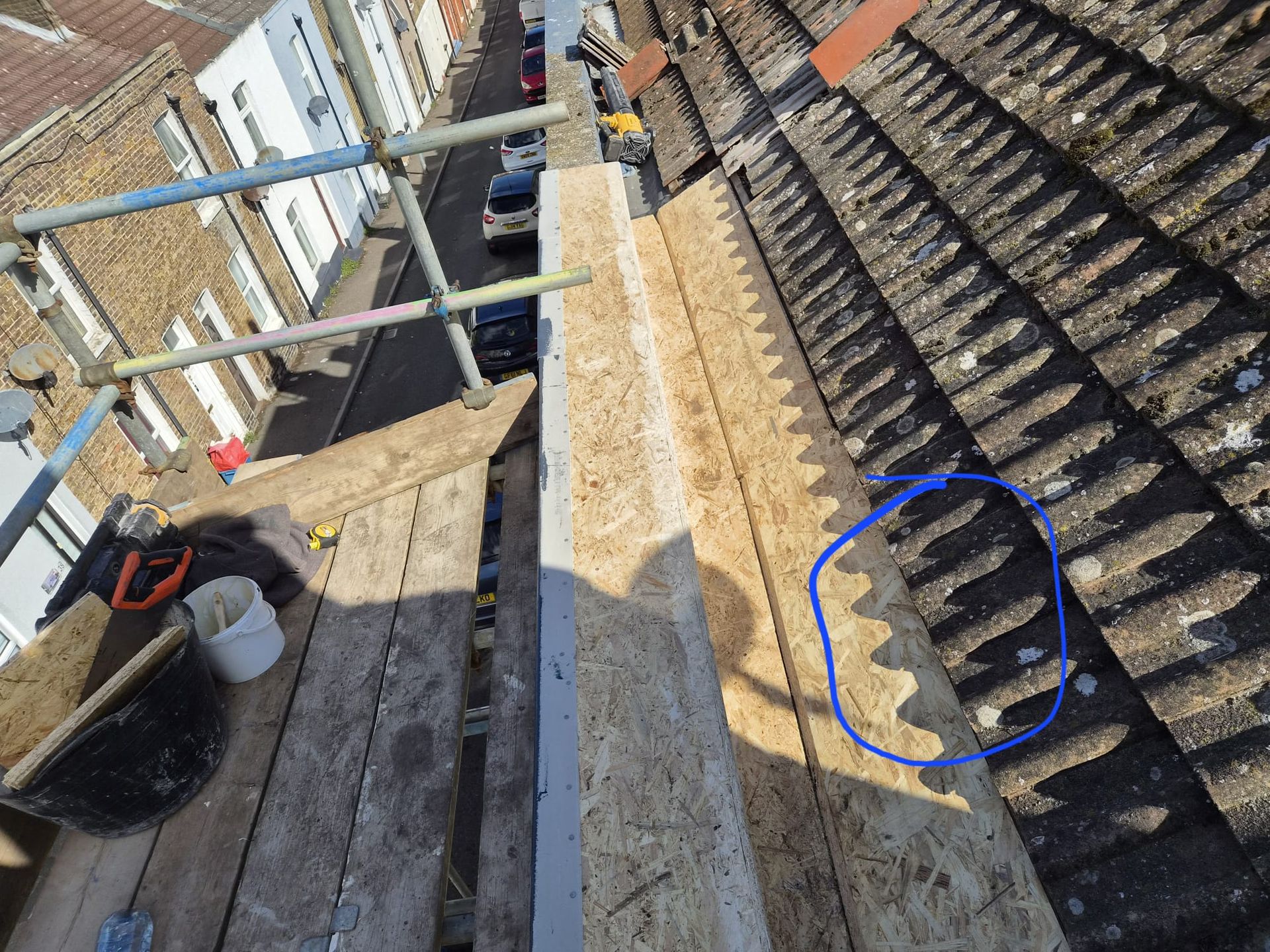 Roof repair work with scaffolding beside a pitched tiled roof, blue circle marking a damaged roof tile area.