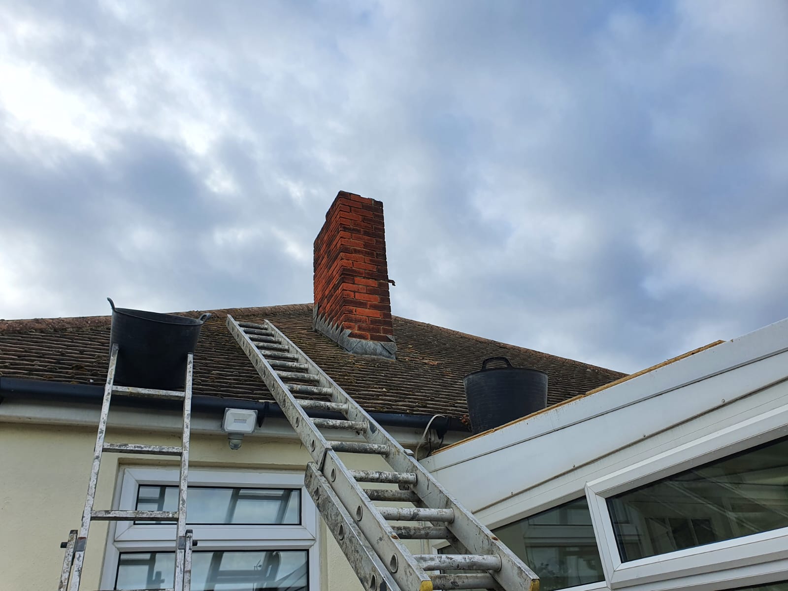 Residential roof with red chimney and ladder under cloudy sky.