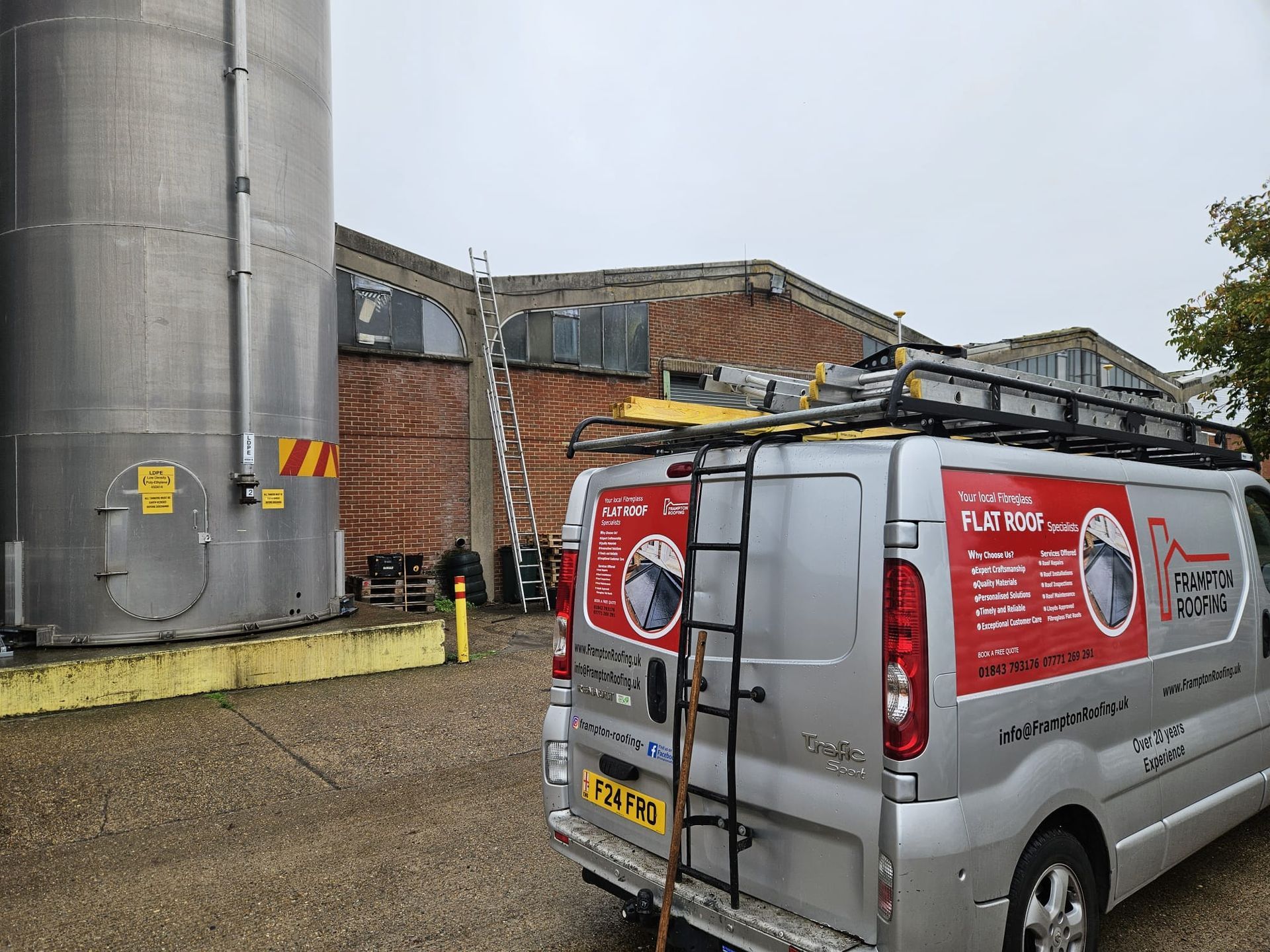 Silver van with ladder and signage, parked next to a large metal tank and brick building.