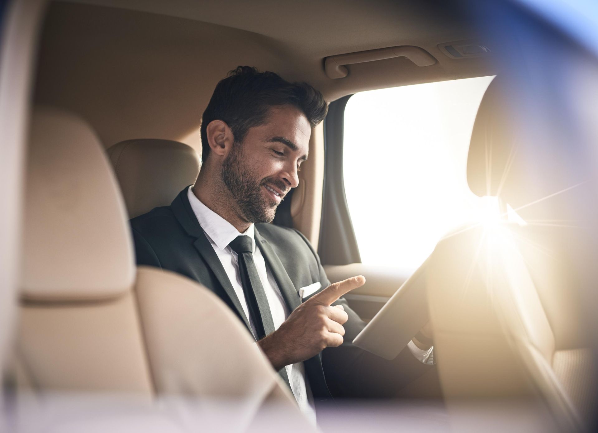 Man in a suit smiling and using a tablet in a sunlit car interior