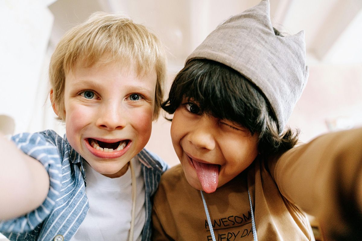 Two smiling children taking a close-up selfie indoors, one sticking out their tongue.