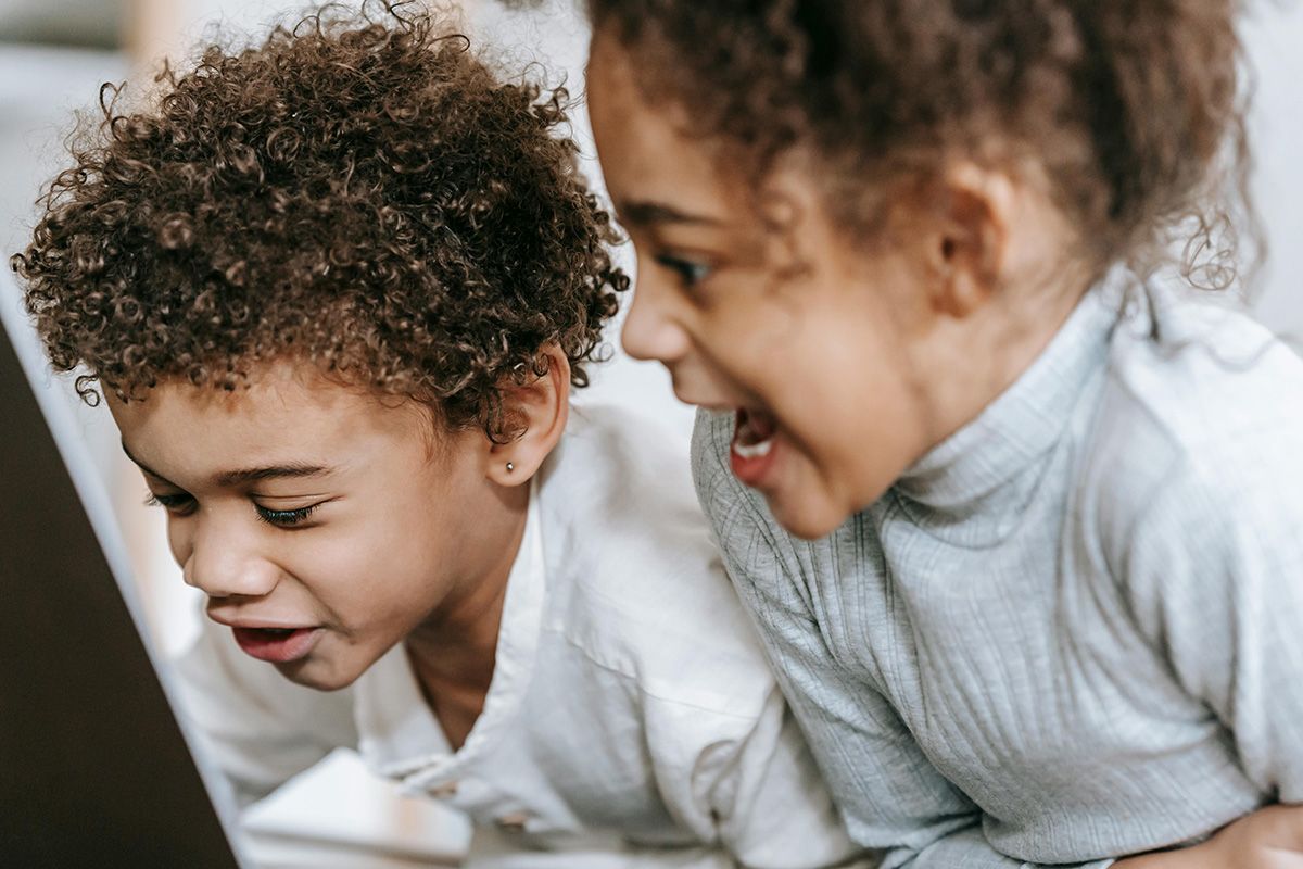 Two children in white sweaters lean close together, smiling and looking down at something off-screen