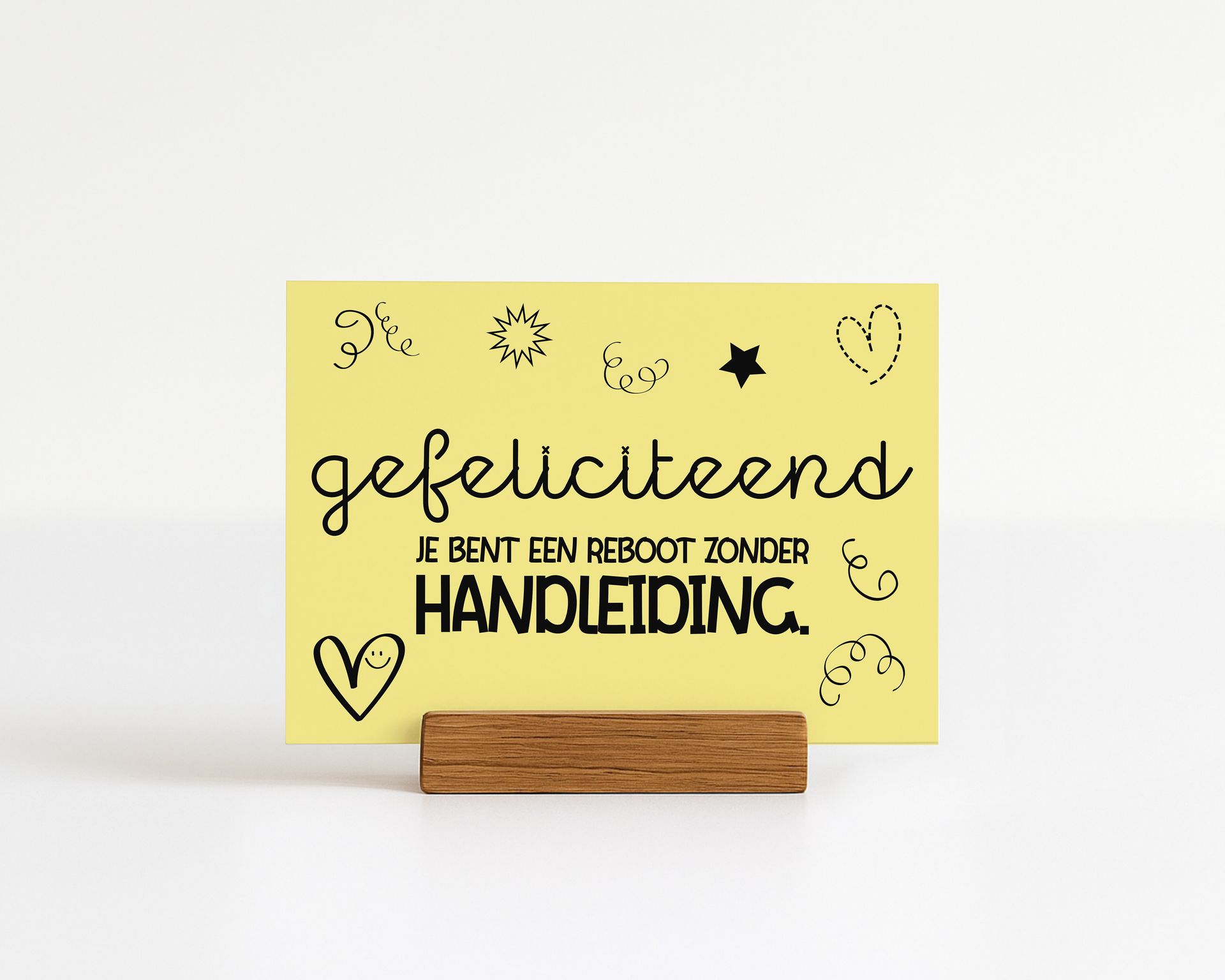 Yellow card with Dutch text "Gefeliciteerd" and "Je bent een reboot zonder handleiding" in wooden holder.