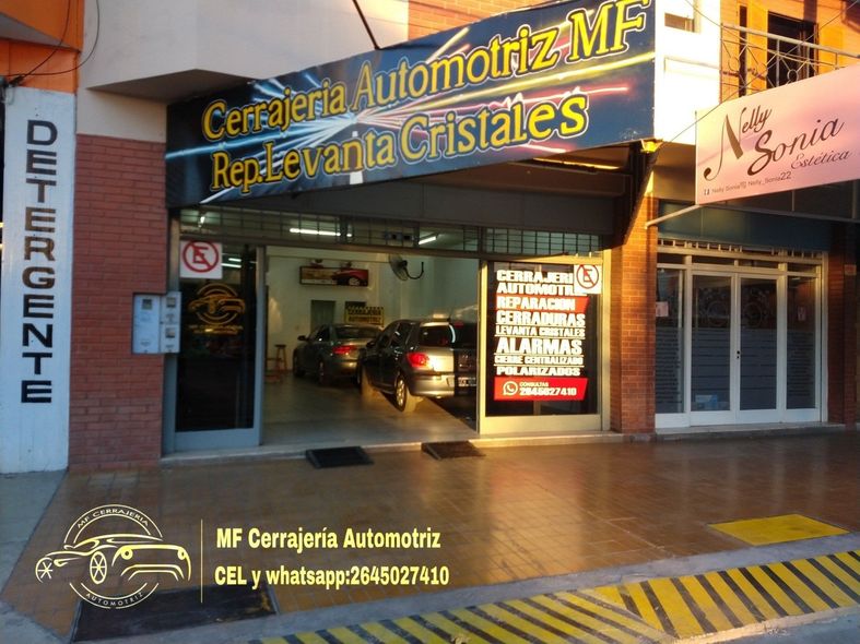 Entrada de taller mecánico con letrero iluminado, garaje abierto y bordillo amarillo de precaución al anochecer.