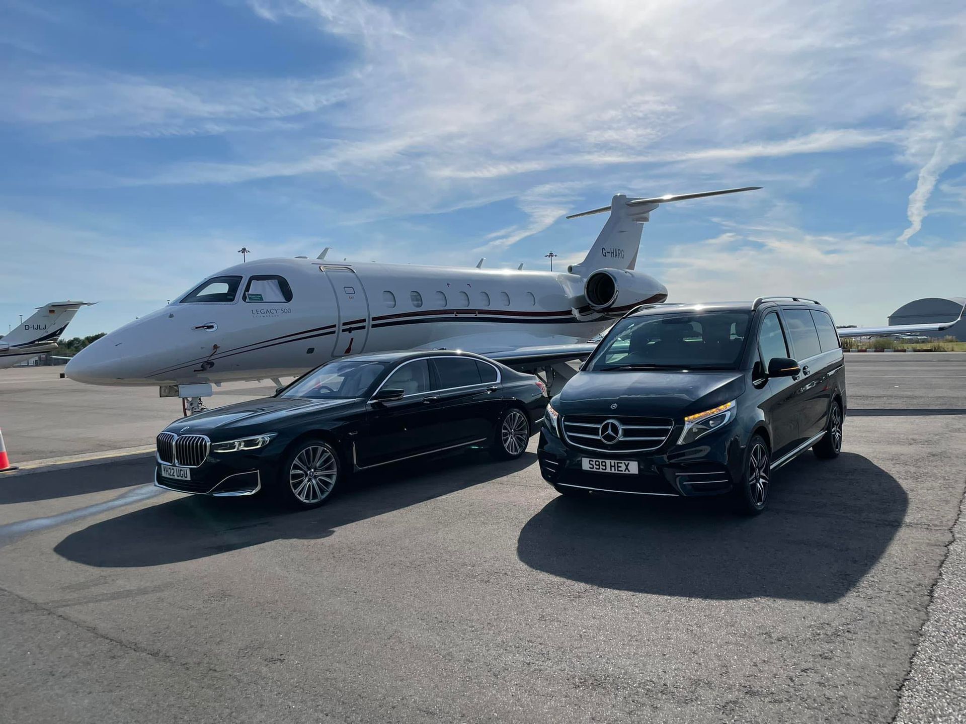 Black sedan and black van parked on an airport tarmac beside a private jet under a blue sky