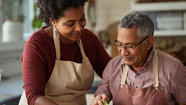 Two people in aprons preparing food together in a kitchen, smiling and focused on a cutting board