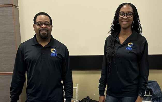 Two people standing indoors in black polos with a blue-and-yellow logo, smiling at the camera.