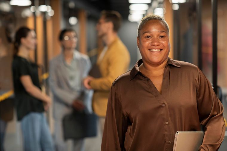 Smiling person in a brown shirt holding a folder in a hallway with people blurred in the background
