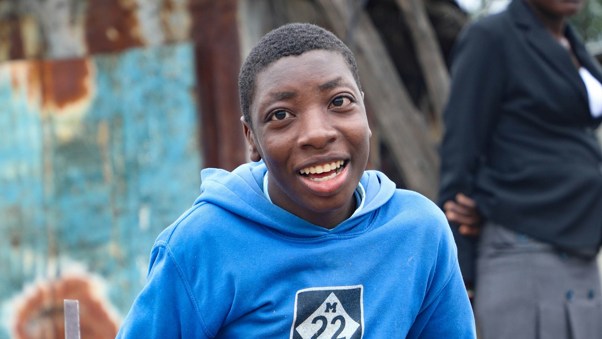 Smiling person in a blue hoodie standing outdoors with a weathered wall behind them