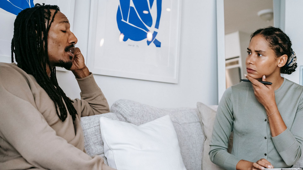 Two people sitting on a couch, facing each other in conversation in a bright room with a white wall hanging.