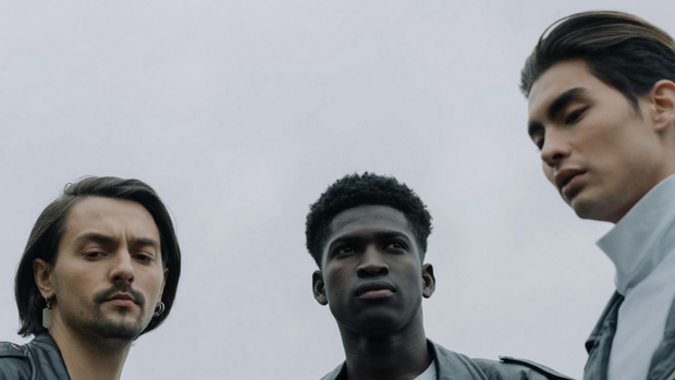Three serious-looking people against a cloudy sky, one in a white coat looking down