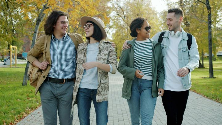 Four friends walking and chatting on a tree-lined path in a park, dressed casually in light jackets.