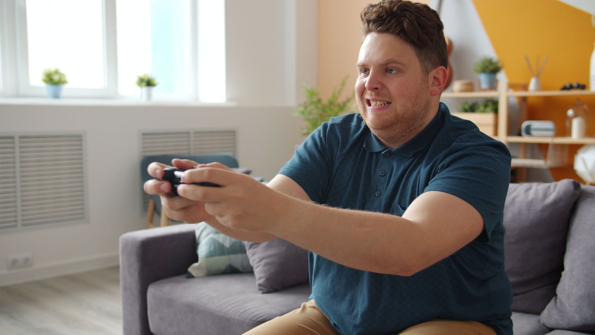 Man on a gray couch smiling while holding a video game controller in a bright living room