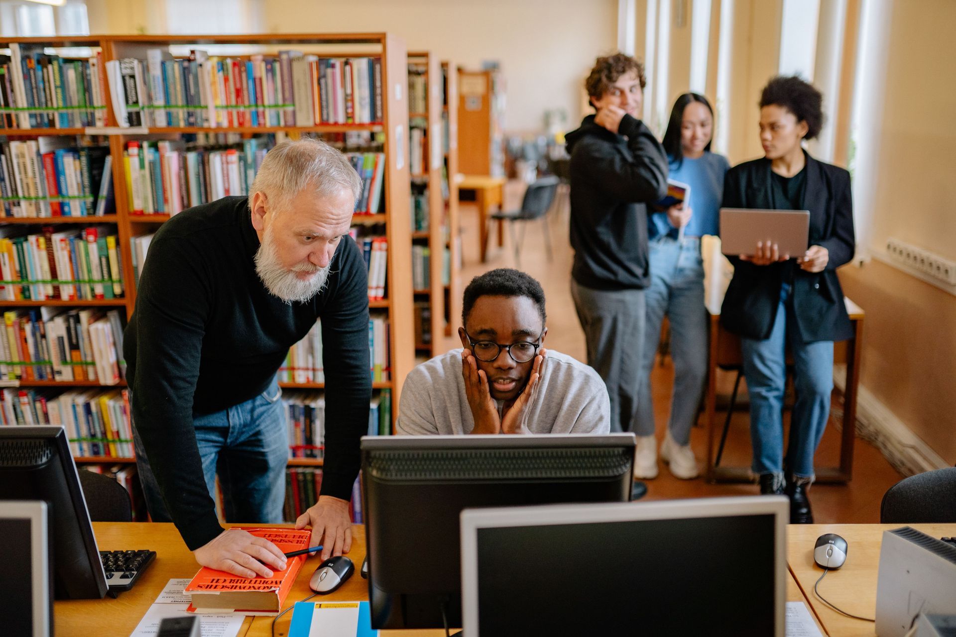 People collaborating at computers in a library, with bookshelves and two others talking in the background.