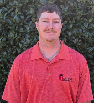 Man in a red polo shirt standing in front of green foliage, smiling at the camera.