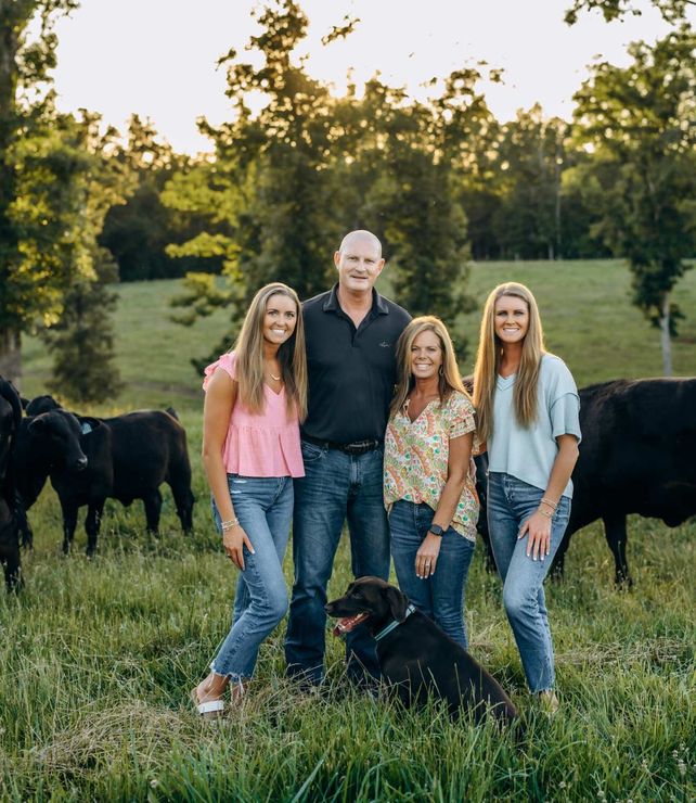 Family standing in a grassy field at sunset with two black dogs and cows in the background