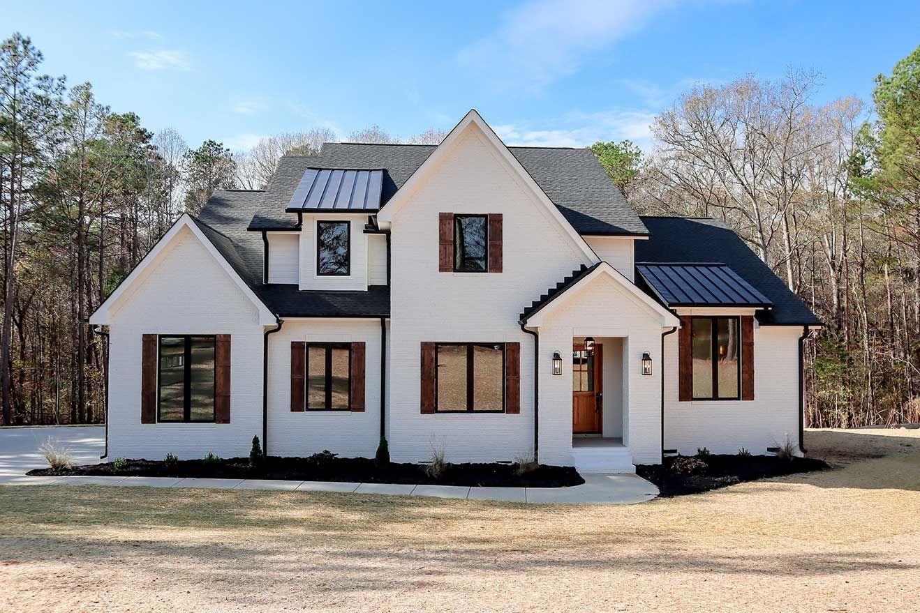 White two-story house with black roof accents and dormer windows in a wooded setting
