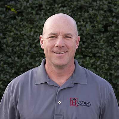 Smiling bald man in a gray polo shirt standing outdoors before green foliage