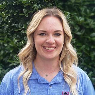 Smiling blonde woman in a blue shirt standing outdoors against green foliage