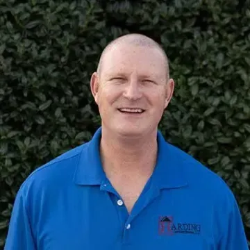 Smiling bald man in a blue polo shirt standing in front of a leafy green hedge
