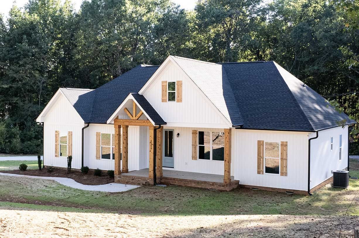 White house with dark roof and front porch on a grassy lot, backed by trees