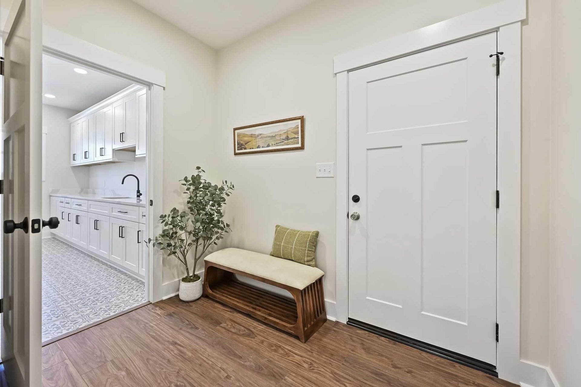 Bright entryway with white door, bench, potted plant, and open view into a kitchen