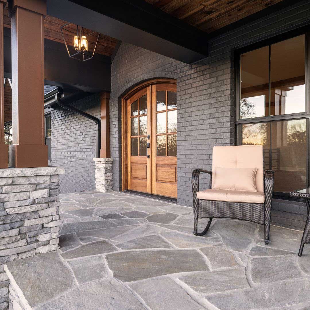 Stone-front porch with arched wooden doors, black trim, and a tan chair beside a window