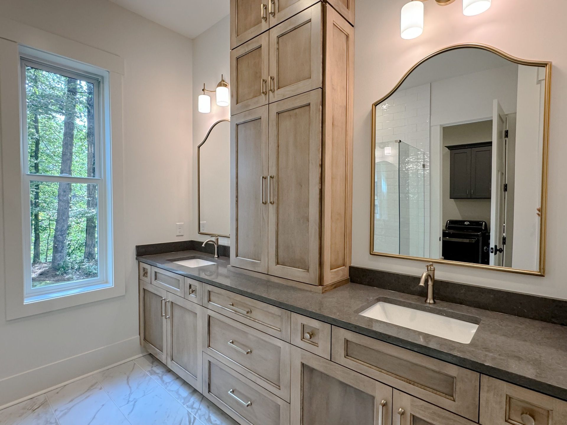 Bright bathroom vanity with double sinks, large arched mirror, and window beside a built-in cabinet.