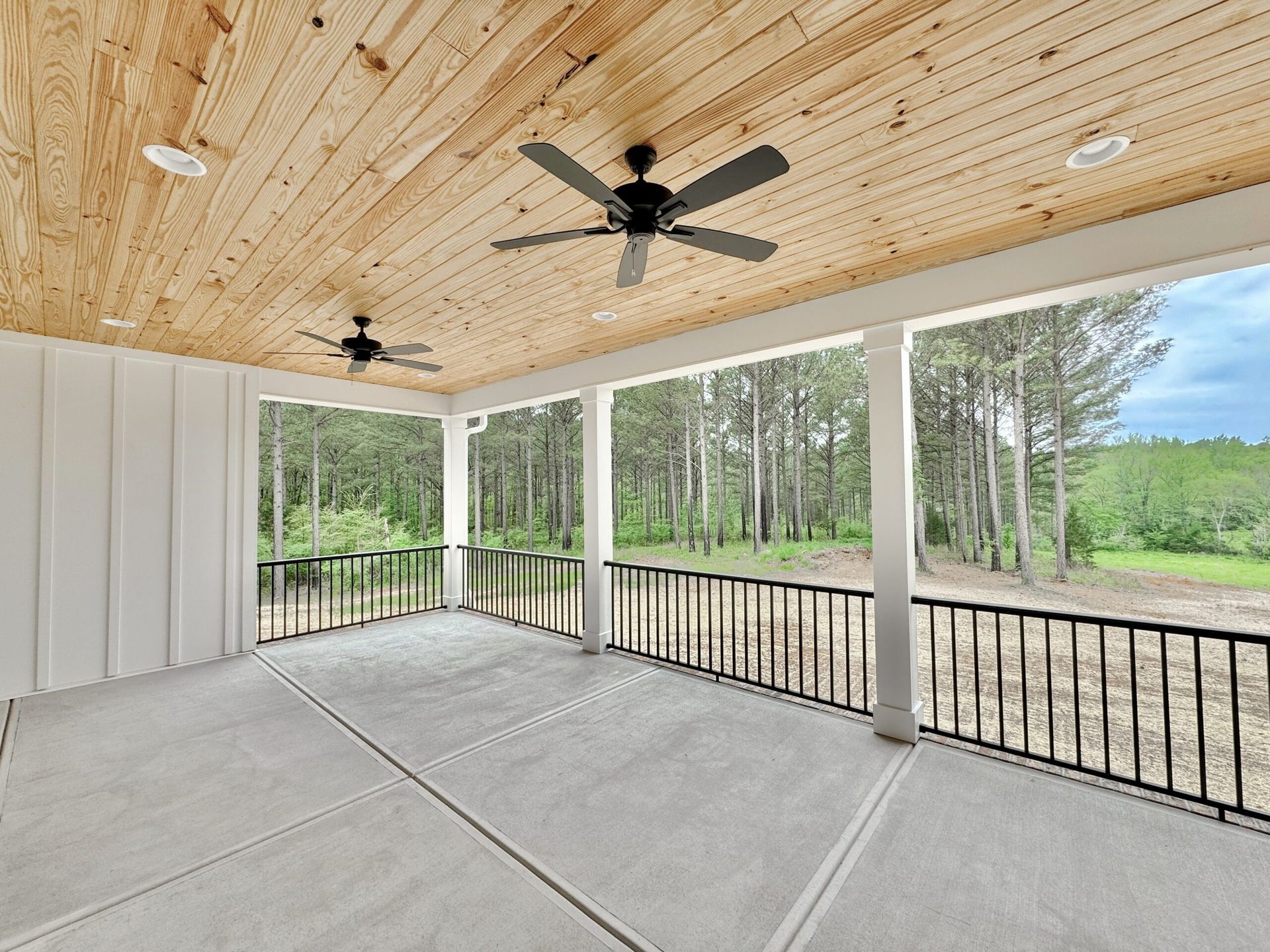 Covered patio with wood ceiling, black fans, and open railing overlooking a grassy yard and trees