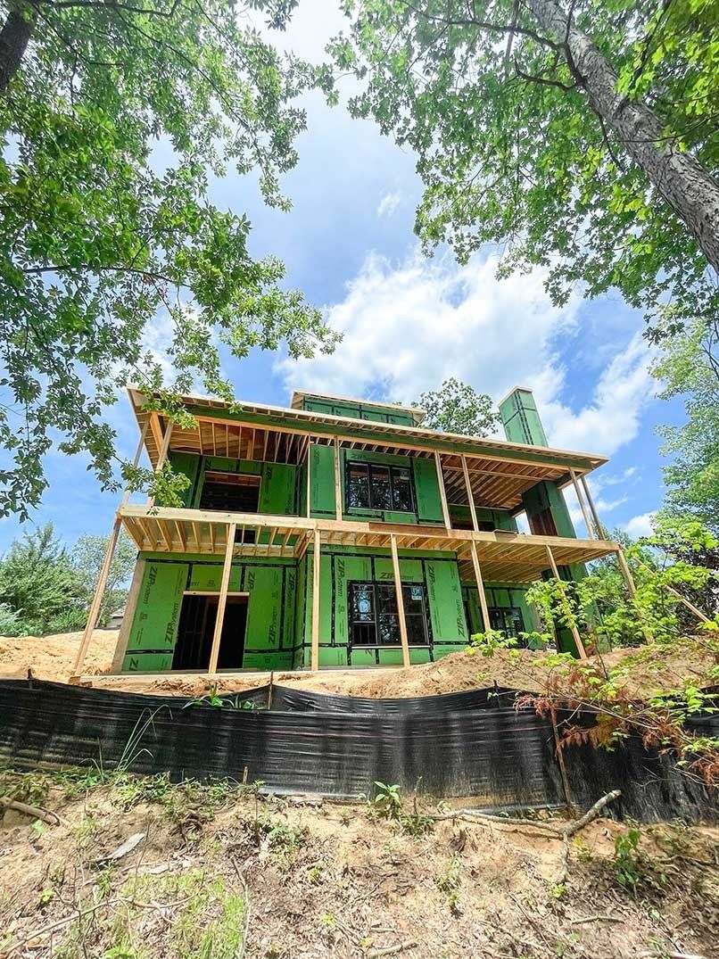 Two-story green house under construction on a hillside, framed by trees and blue sky