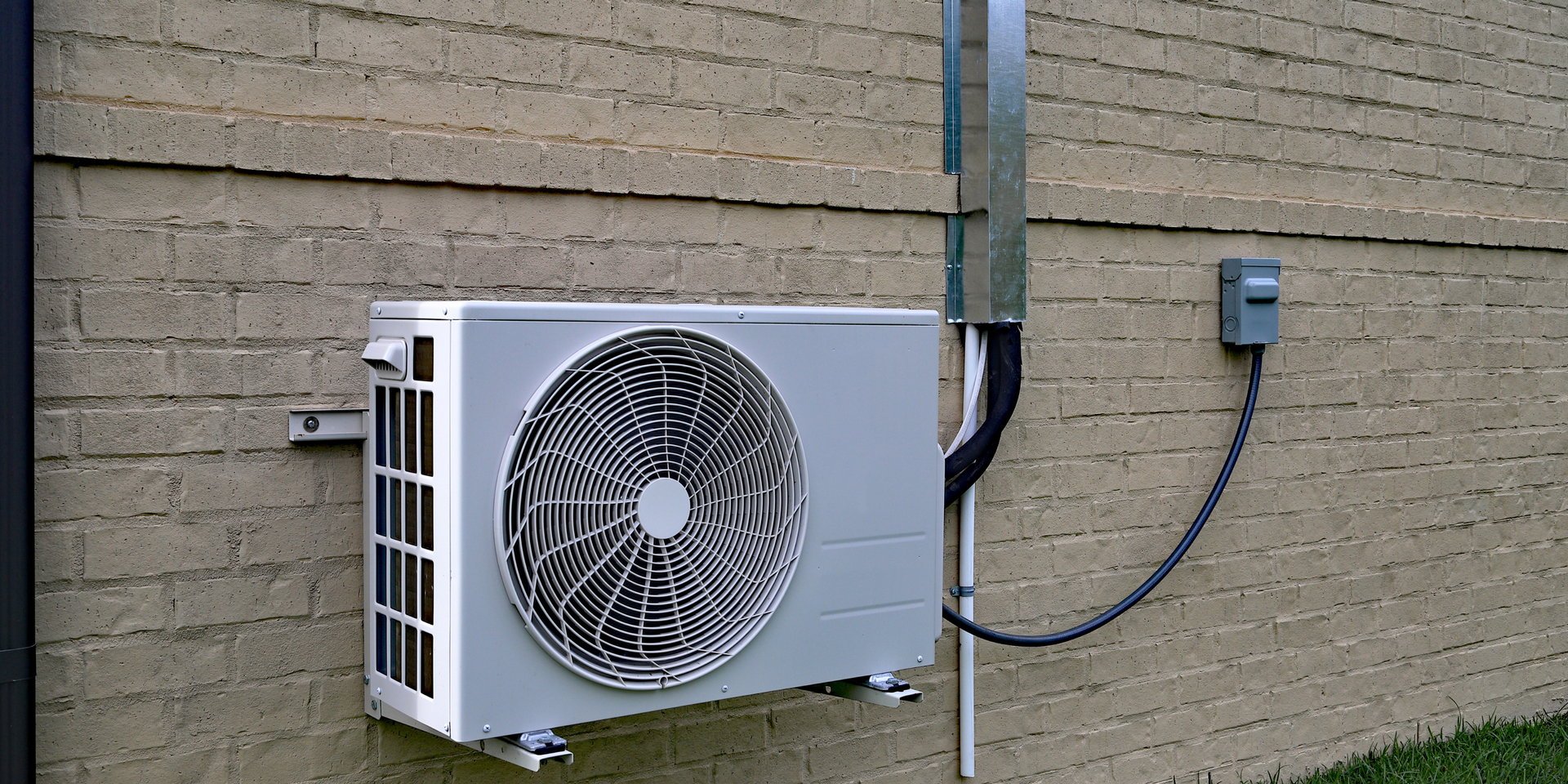 A white mini-split air conditioner condenser mounted on a tan brick wall next to a power disconnect box and conduit.