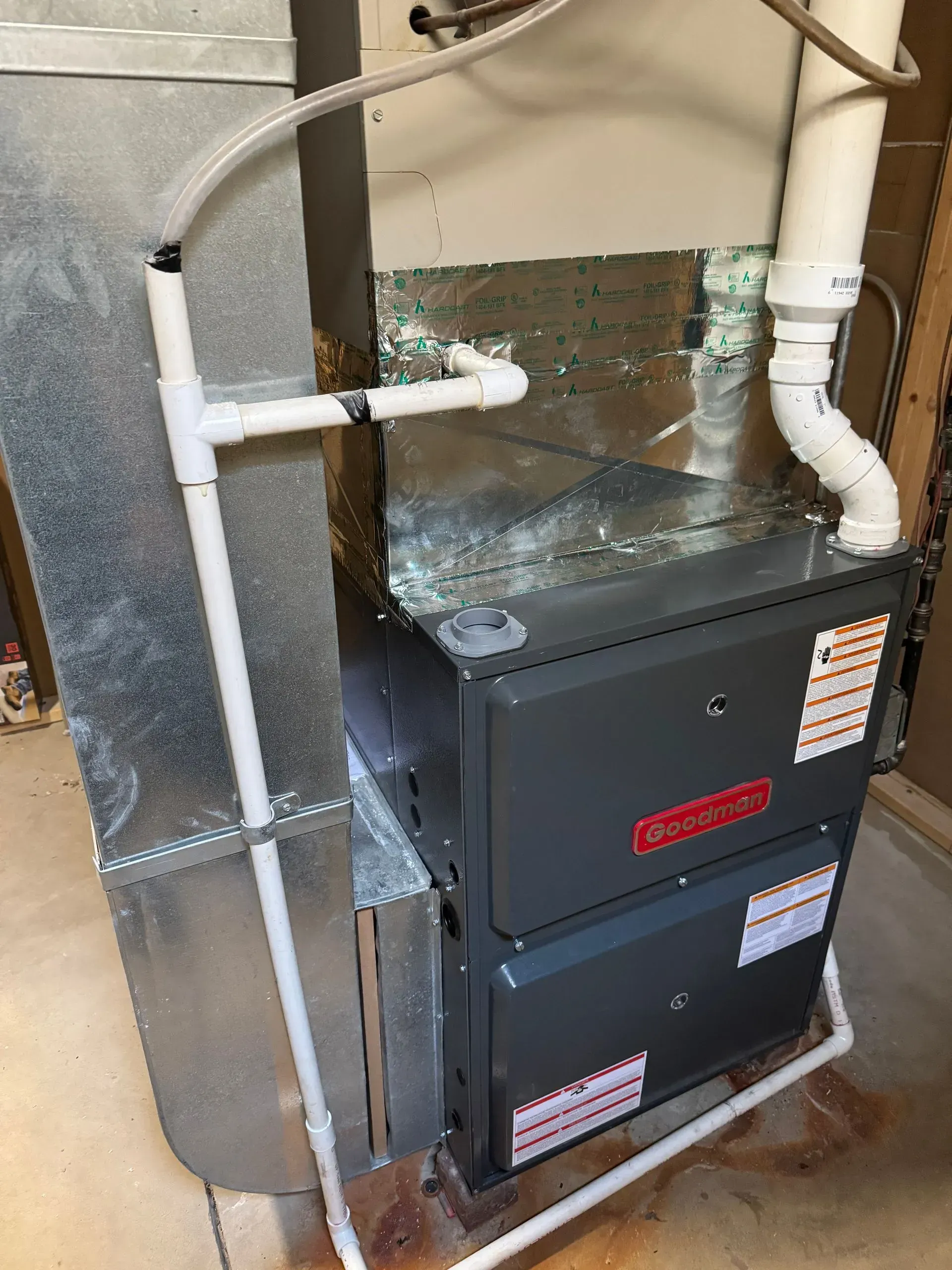 A Goodman furnace in a utility room with white PVC pipes connected to it and metal ductwork.