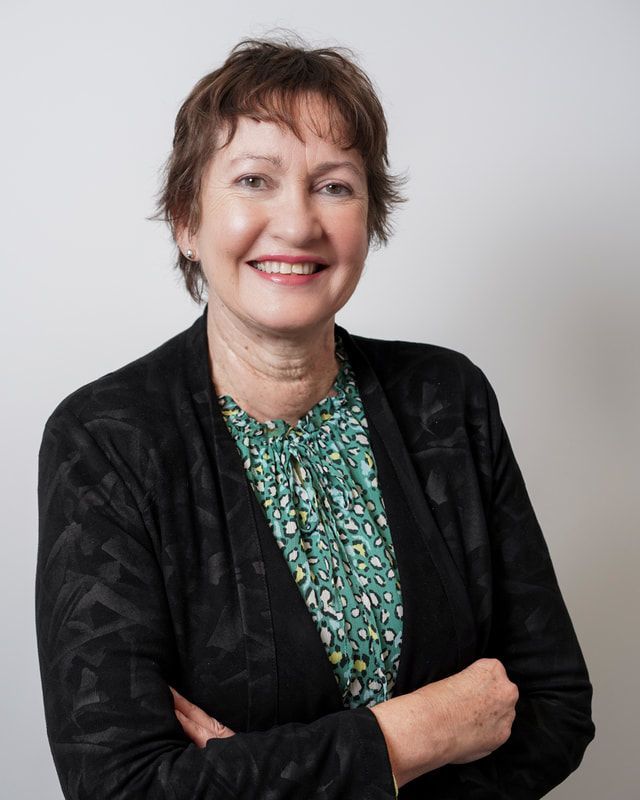 A woman wearing a black jacket and a green shirt is smiling with her arms crossed.