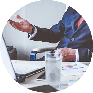 A man in a suit and tie is sitting at a desk with a laptop and a glass of water.