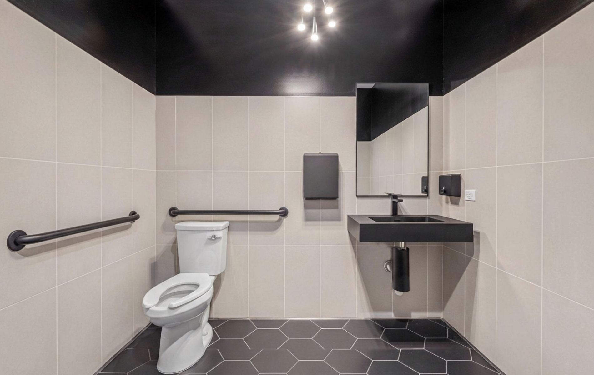Modern accessible restroom with toilet, grab bars, black sink, mirror, and tiled walls and floor