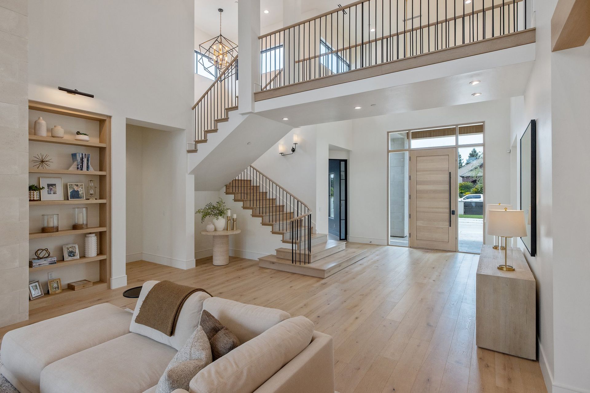 Bright modern living room with open staircase, white walls, wood floors, and tall windows.