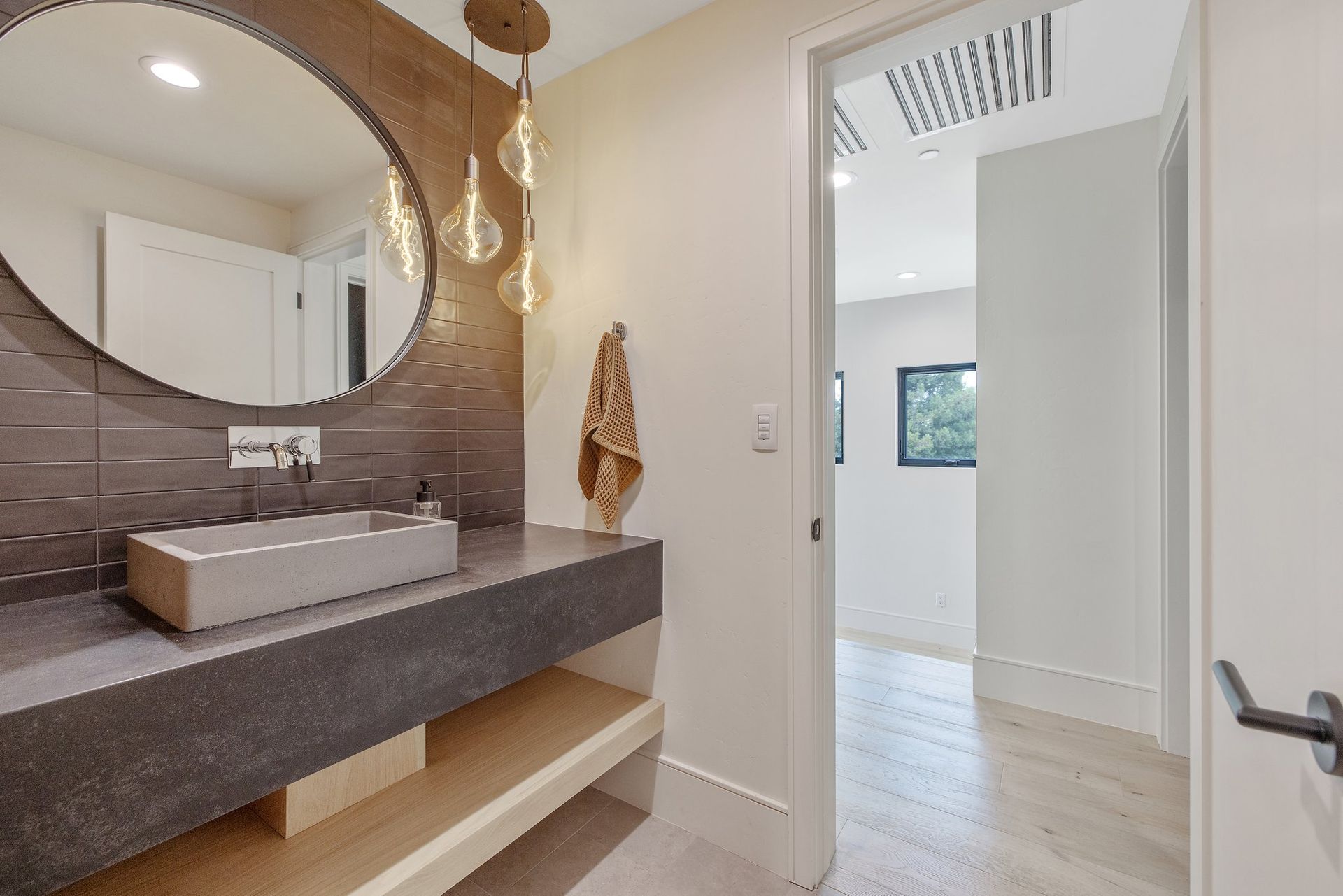 Modern bathroom vanity with dark countertop, round mirror, pendant lights, and doorway to a bright room