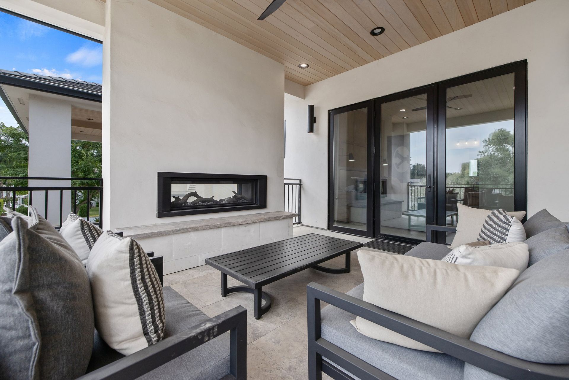 Covered patio with gray seating, coffee table, and a wall-mounted fireplace beside glass doors.
