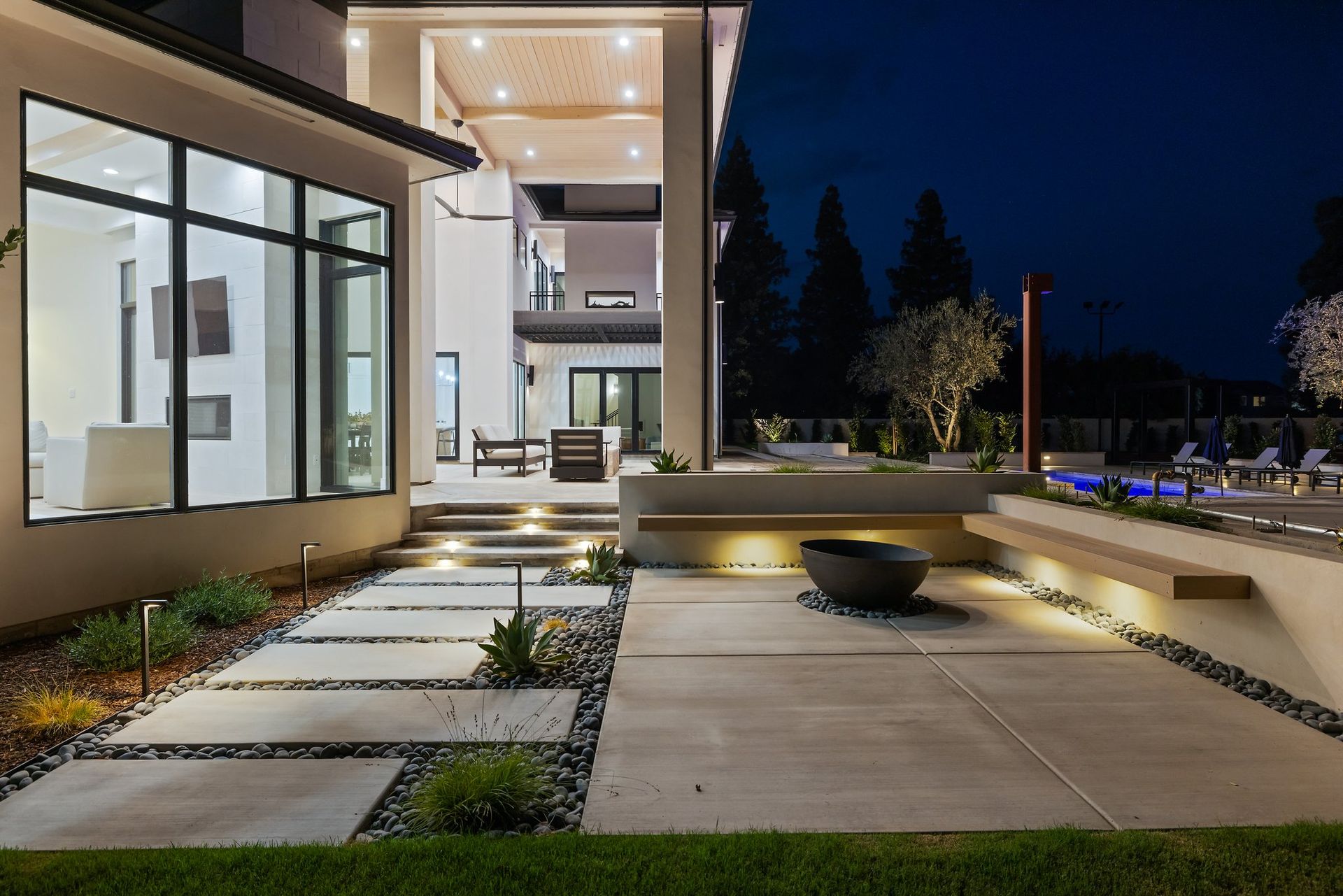 Modern house exterior at night with lit walkway, glass windows, and landscaped patio