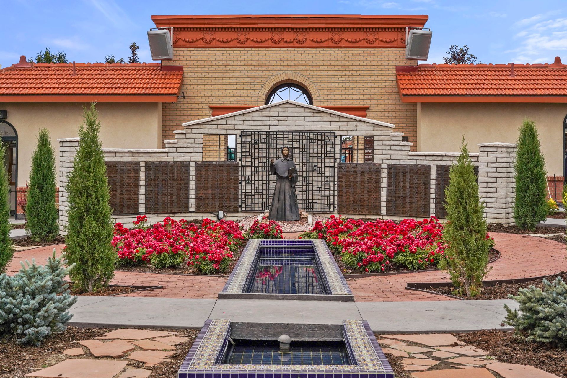 Formal garden with fountains, red flowers, and a gated entrance to a stucco building.