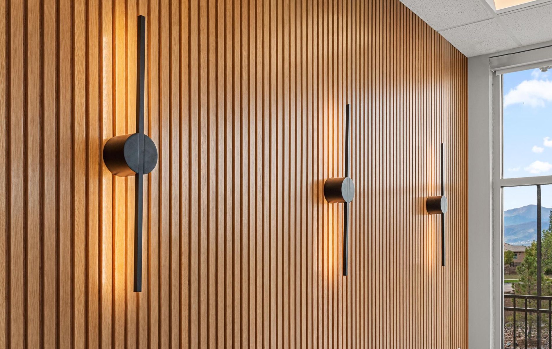 Wood-paneled wall with minimalist black sconces beside a bright window and mountain view