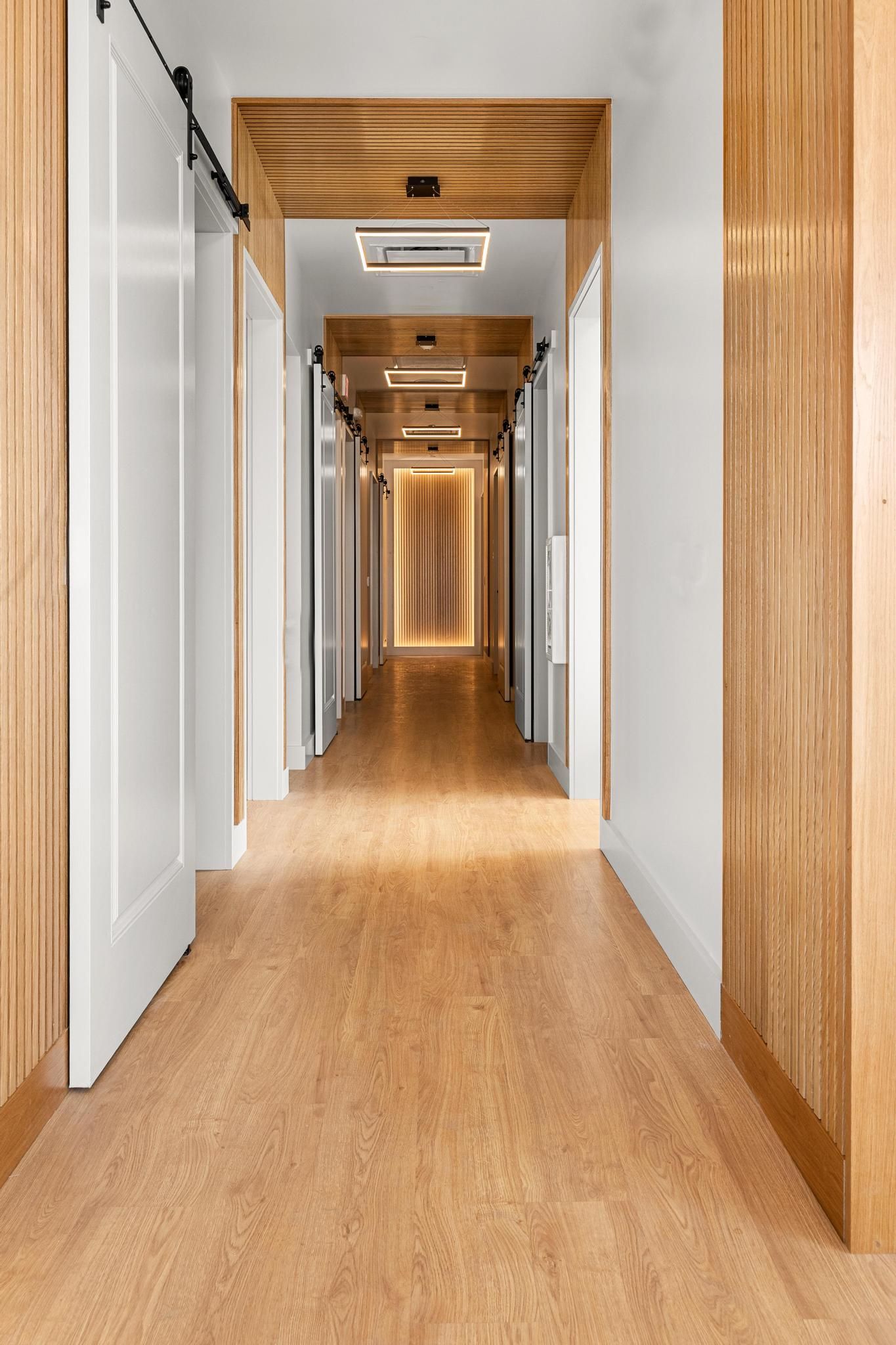 Modern hallway with wood flooring, white walls, and wooden ceiling accents, leading to a bright doorway.