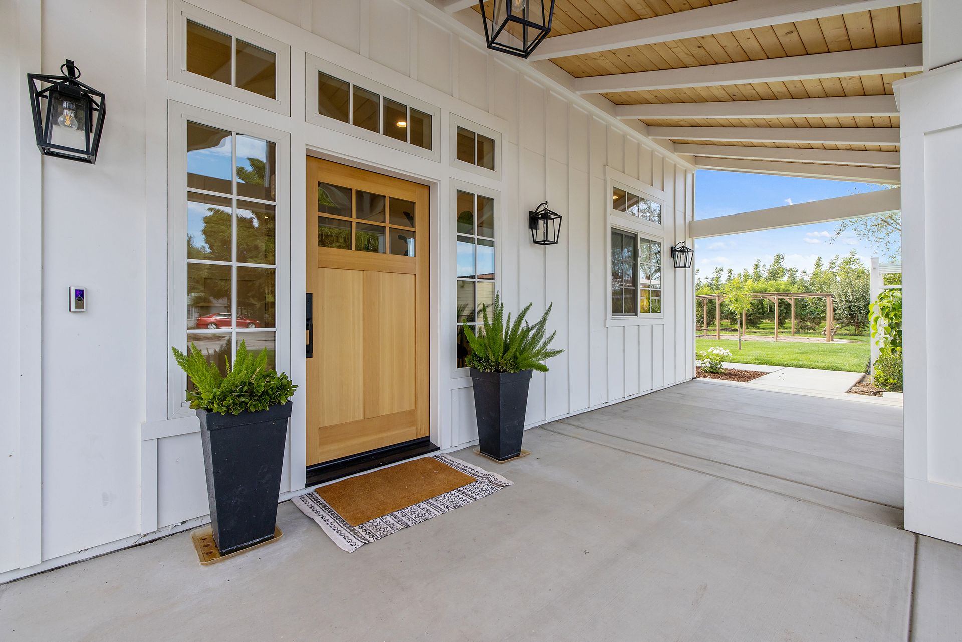Covered front porch with white siding, wooden door, black lanterns, and potted plants leading to a bright walkway