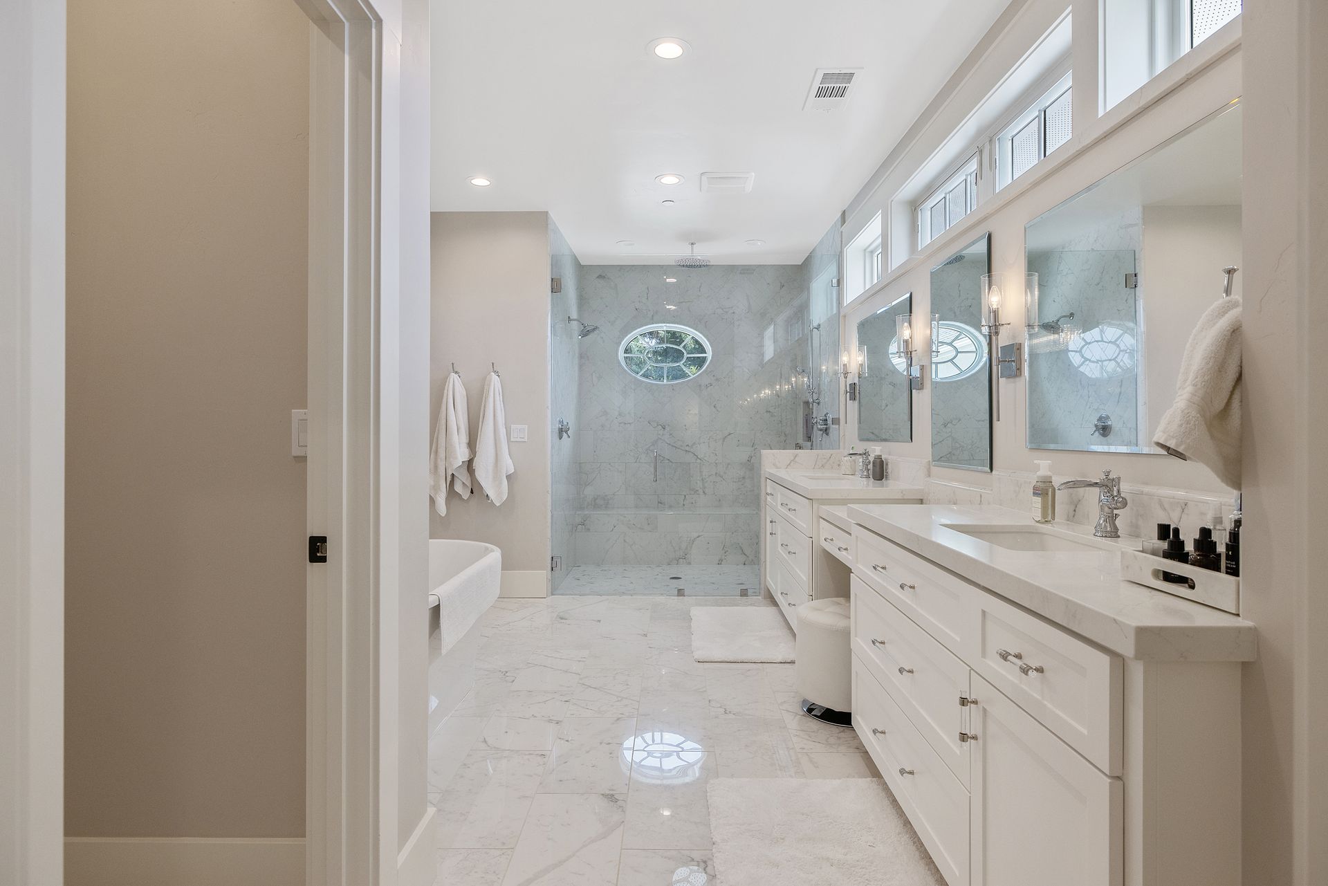 Bright marble bathroom with double vanity, large mirrors, and glass shower under skylights.