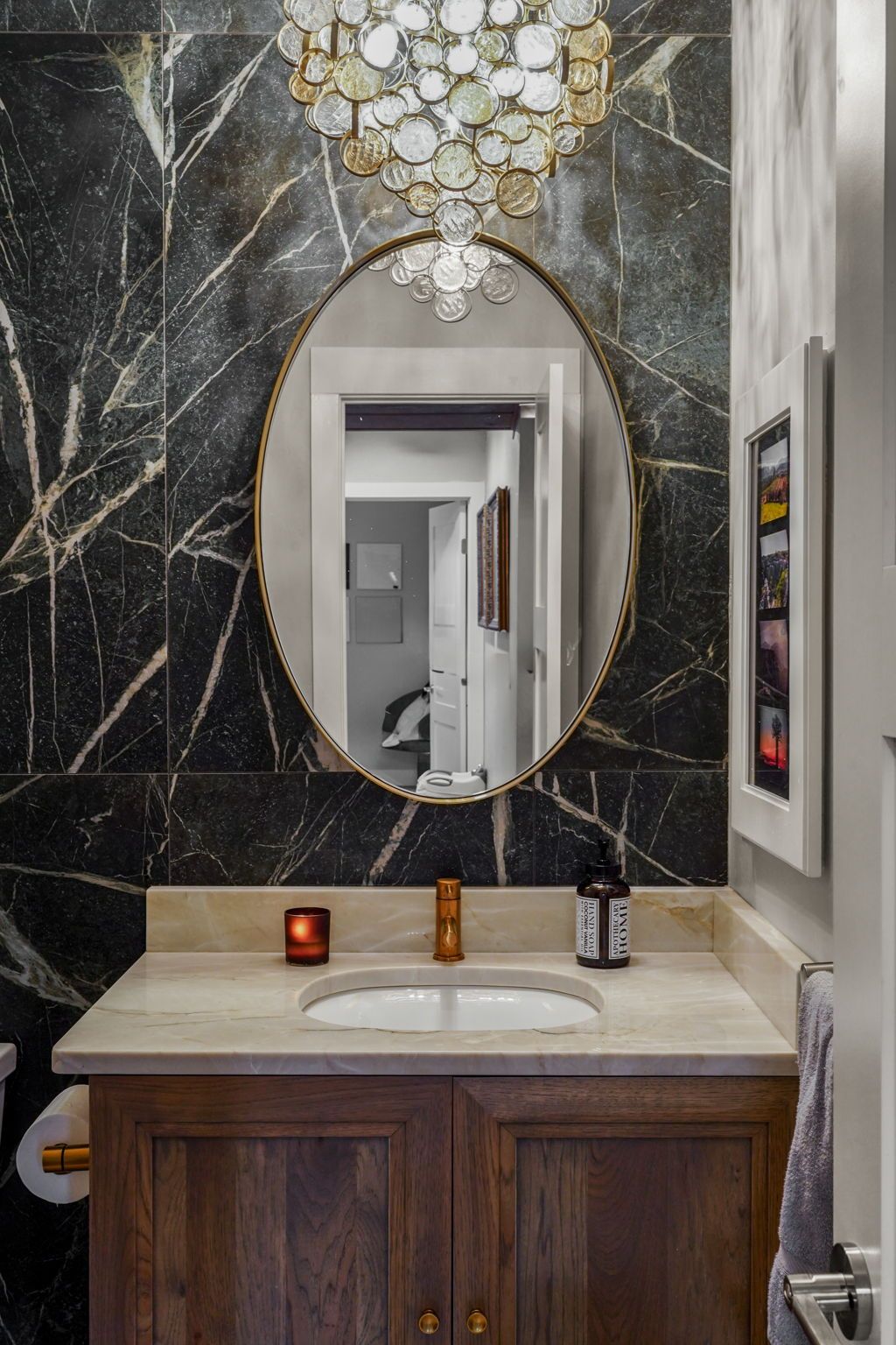 Bathroom vanity with oval mirror, black marble wall, gold chandelier, and wooden cabinet.