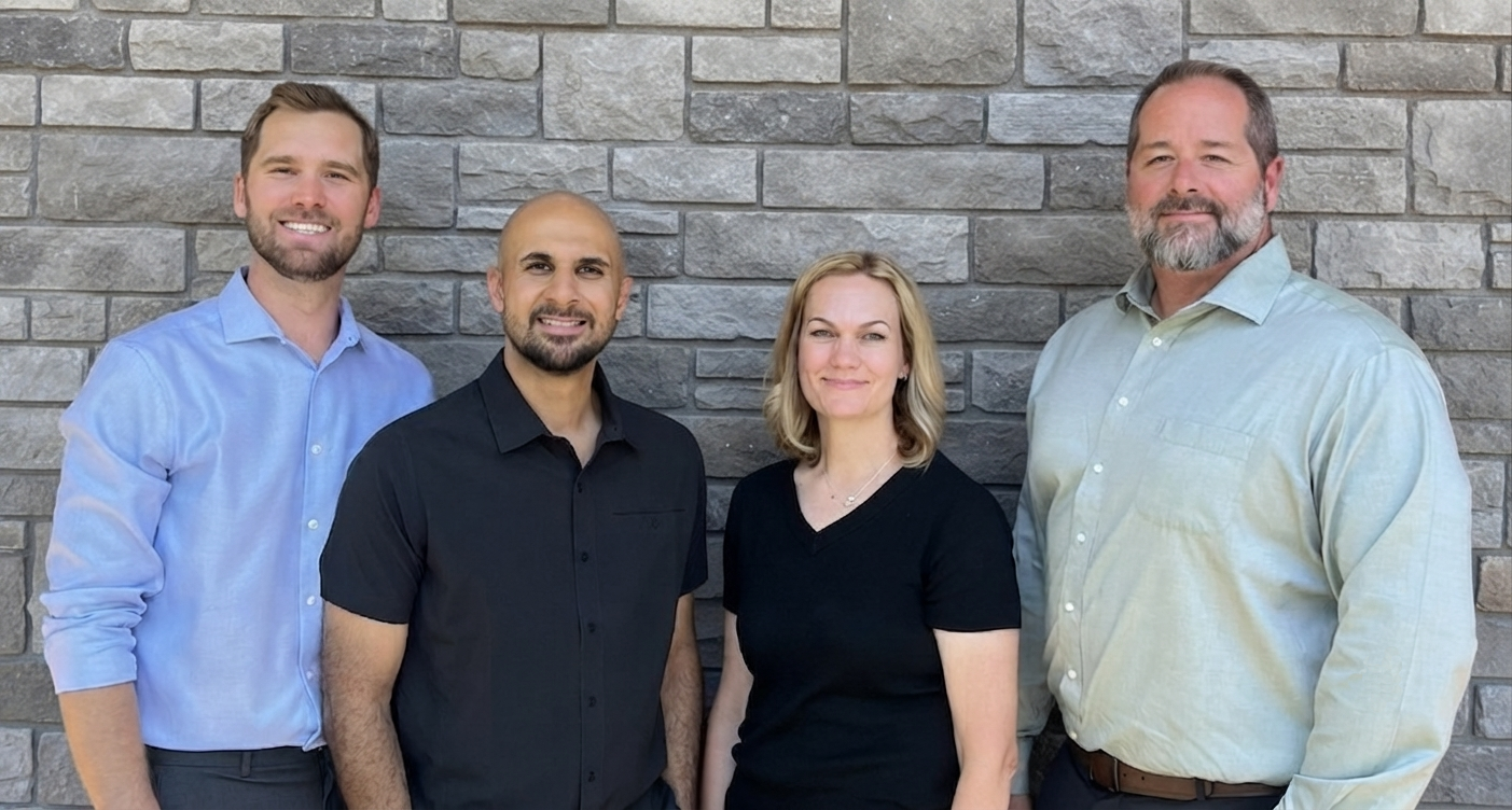 Four people standing in front of a gray stone wall, facing the camera and smiling.