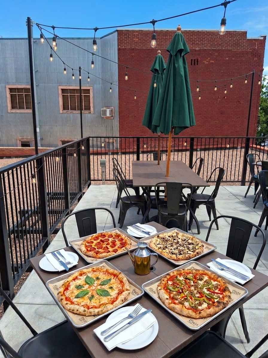 Outdoor rooftop table with three pizzas, a drink, and black chairs under a green umbrella