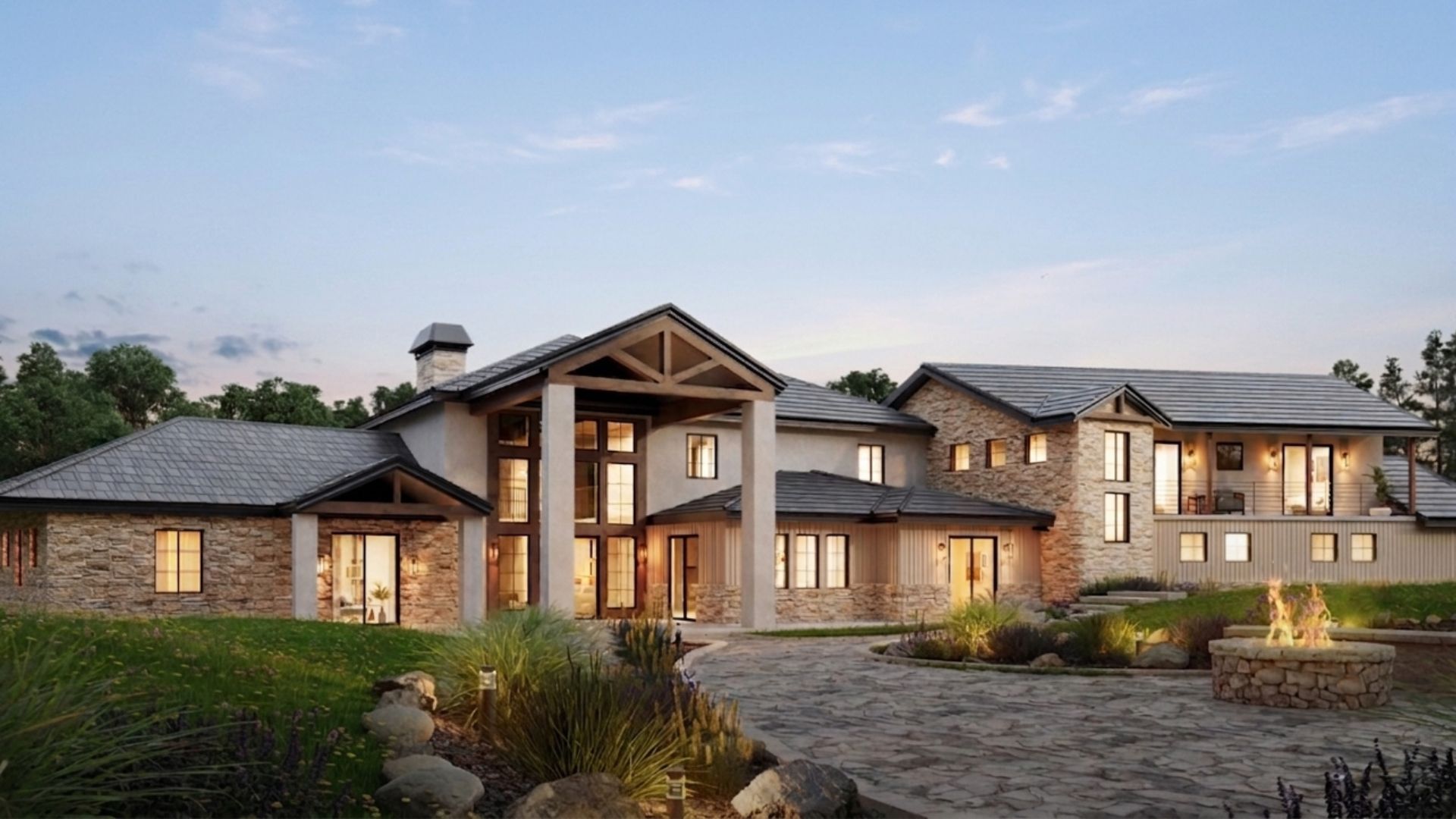 Large stone-and-wood mansion at dusk with lit windows, landscaped garden, and a cobblestone driveway.