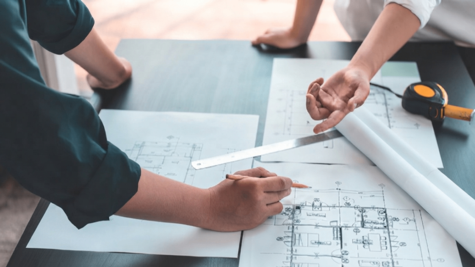 Hands drafting architectural plans on a table with rulers and rolled blueprints